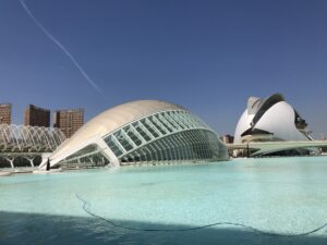 The glass dome of L'Hemisferic in Valencia reflected in a shallow pool, with the white shell of the Palau de les Arts on the right