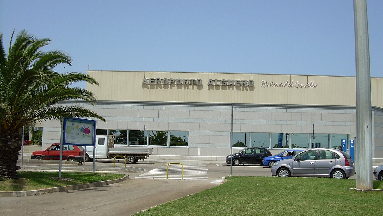 Budova letiště Alghero Riviera del Corallo s palmou a zaparkovanými auty před vchodem