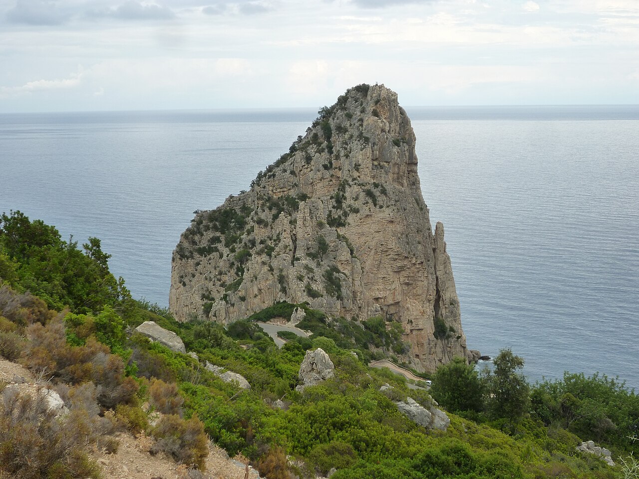 Skalní jehla Pedra Longa u Baunei nad mořem, pod ní klikatá silnice a macchie