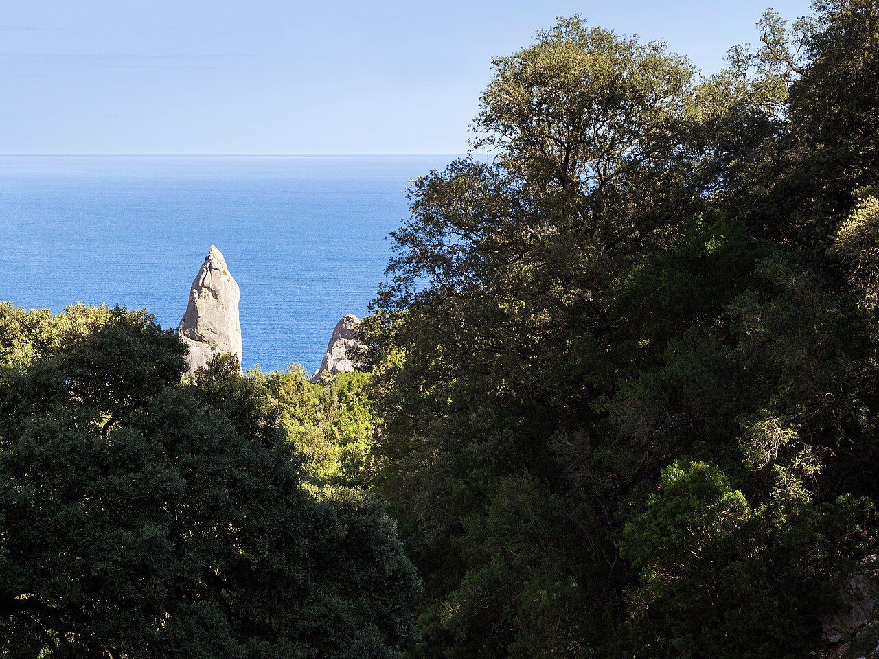 Skalní jehla nad zátokou Cala Goloritzé mezi korunami stromů, pohled ze sestupové stezky k moři