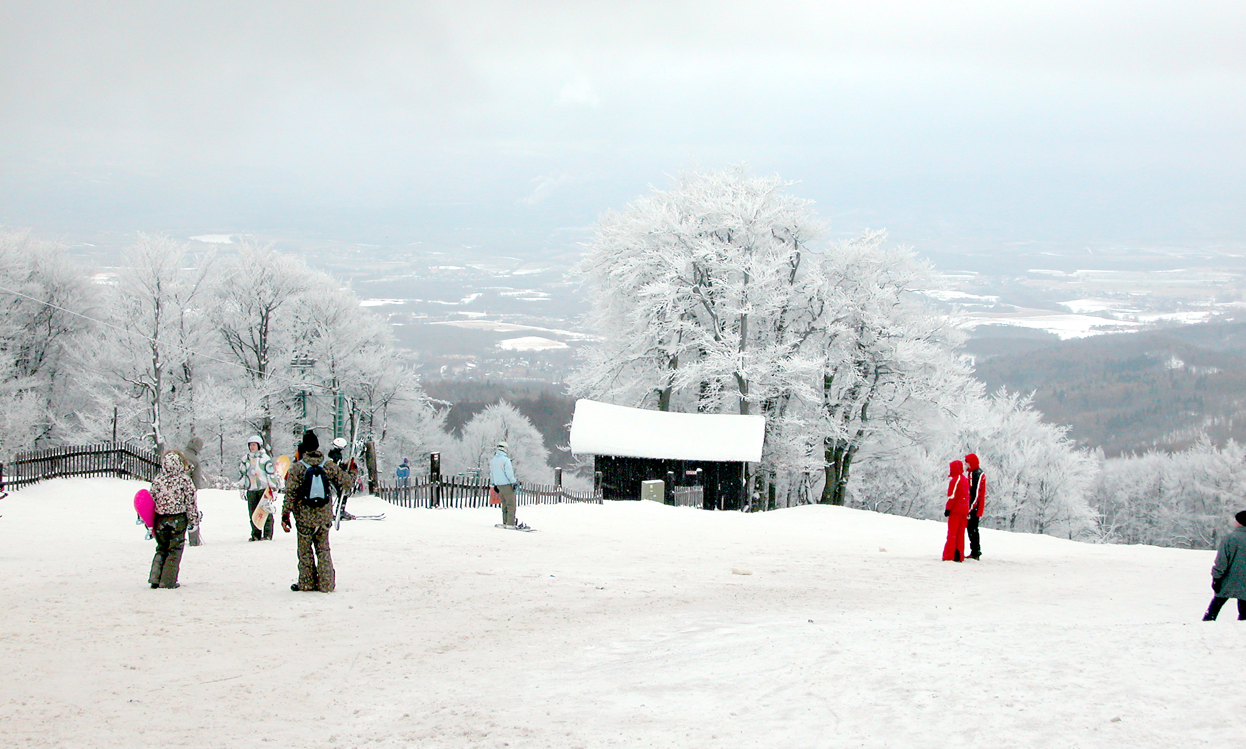 Lyžaři a snowboardisté na zasněženém vrcholu Bouřňáku, kolem stromy obalené námrazou a ve výhledu krajina pod Krušnými horami