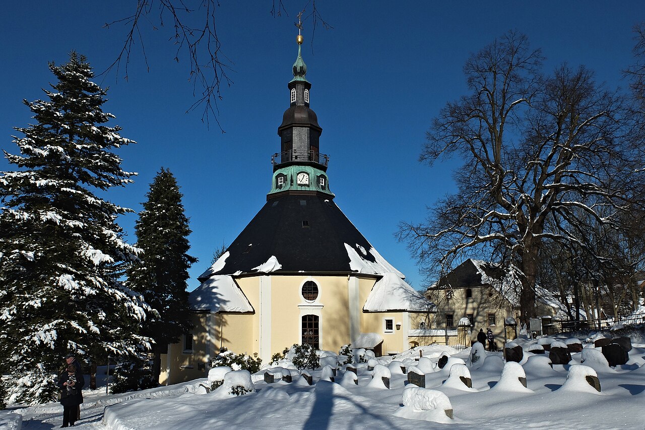 Osmiboký barokní kostel Bergkirche v Seiffenu v zimě, zasněžený hřbitov a modrá obloha