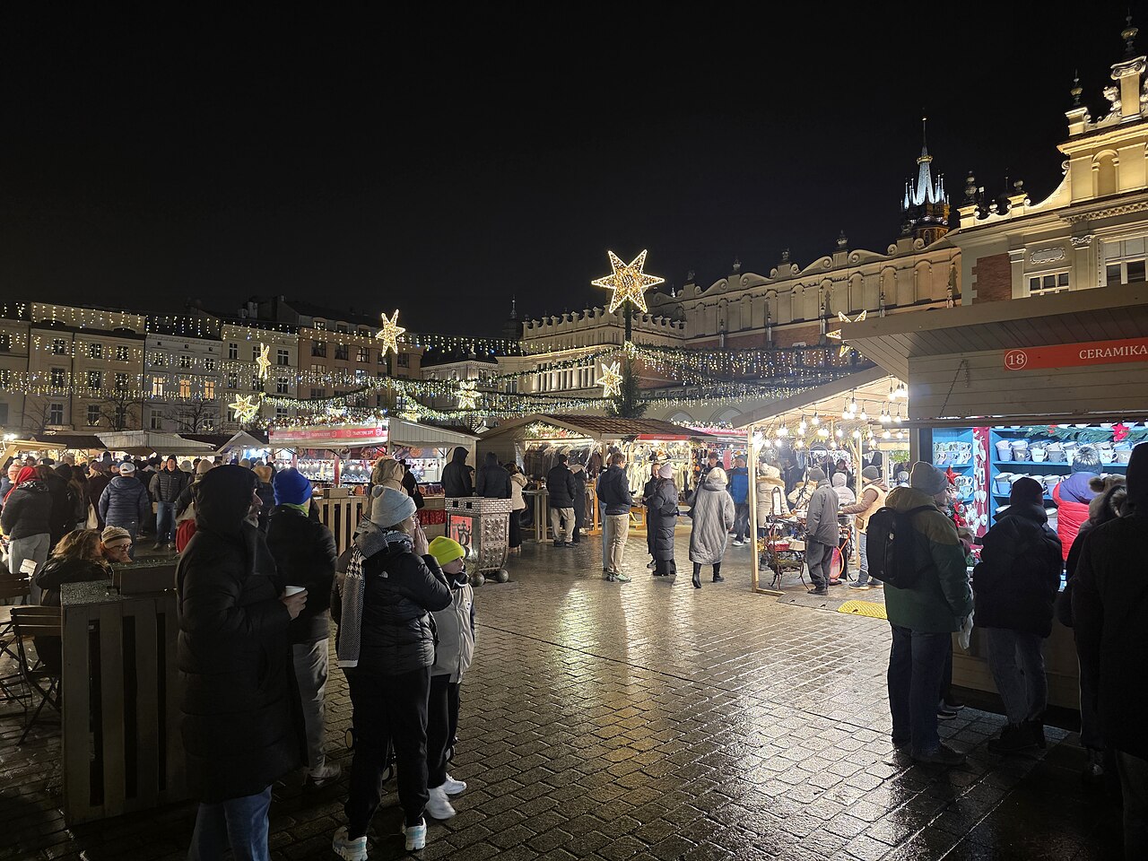 Krakow Christmas market in the evening: lit stalls, glowing stars and people with mulled wine on wet cobbles in front of the Cloth Hall