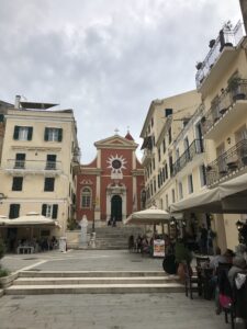 Plaza del casco antiguo de Corfú con una iglesia de fachada roja y cafés