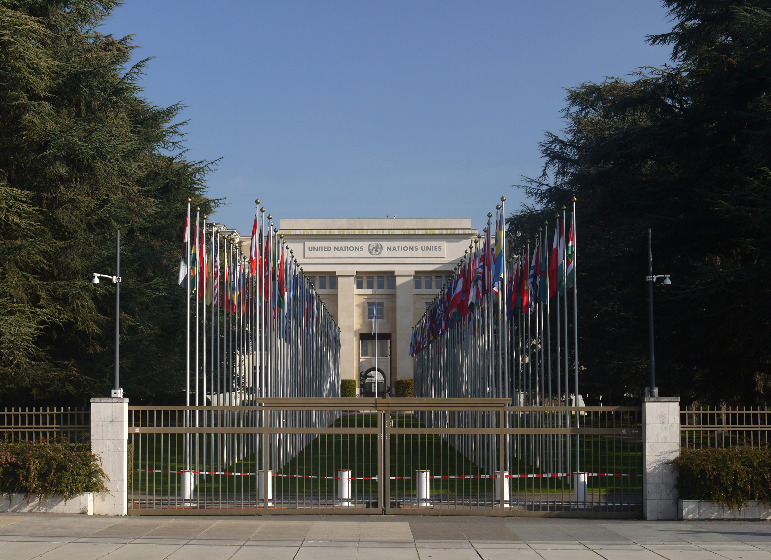 Alej vlajek členských států vedoucí k budově Paláce národů s nápisem United Nations Nations Unies, v popředí zavřená brána