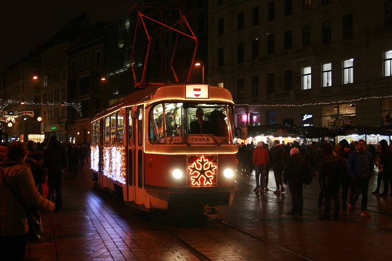 Świątecznie oświetlony brneński tramwaj przejeżdża nocą przez plac Wolności między tłumem a straganami jarmarku bożonarodzeniowego