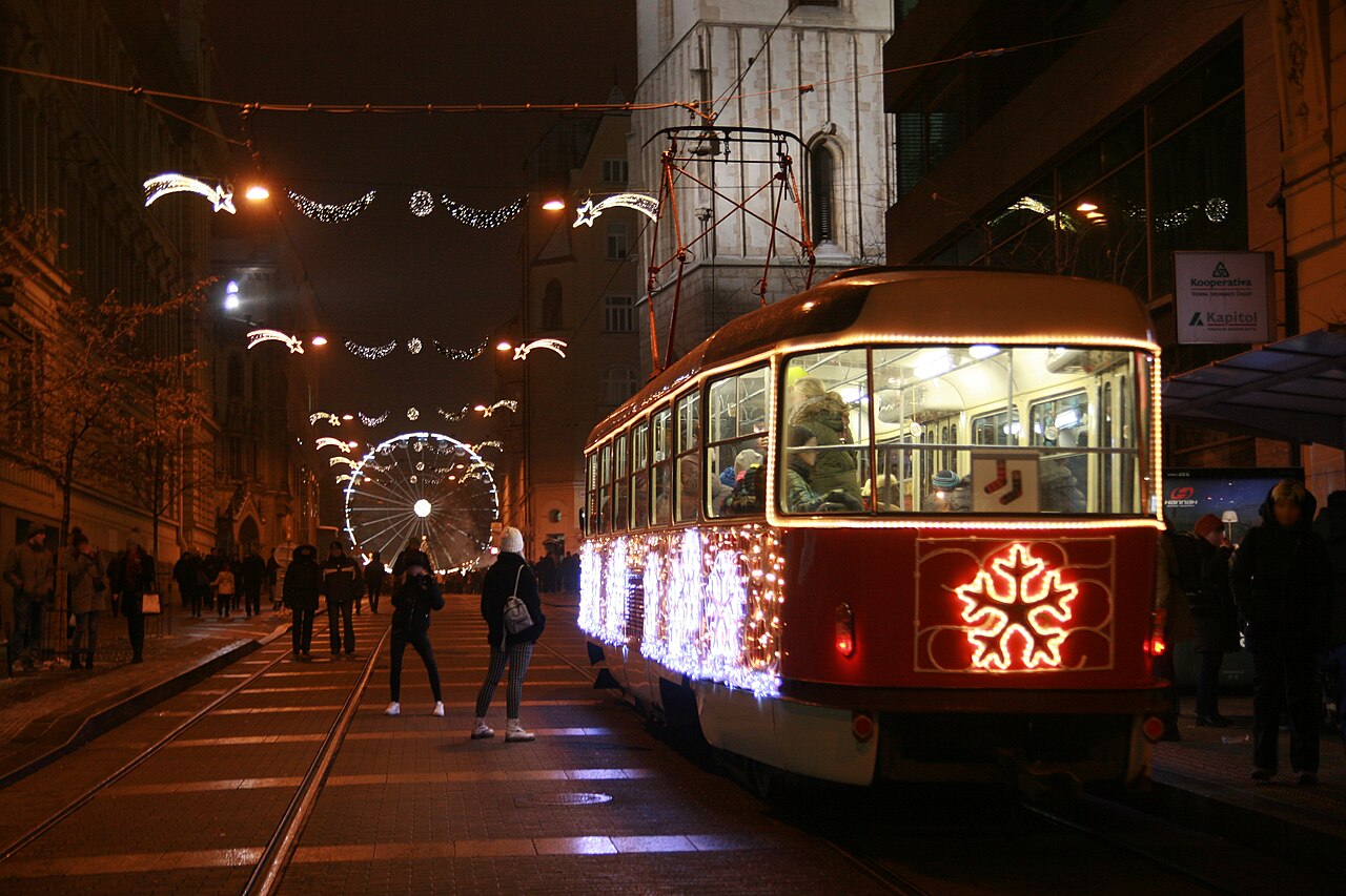 Oświetlony świąteczny tramwaj nocą na ulicy Rašínovej w Brnie, nad ulicą świetlne dekoracje, w tle wieża kościoła św. Jakuba