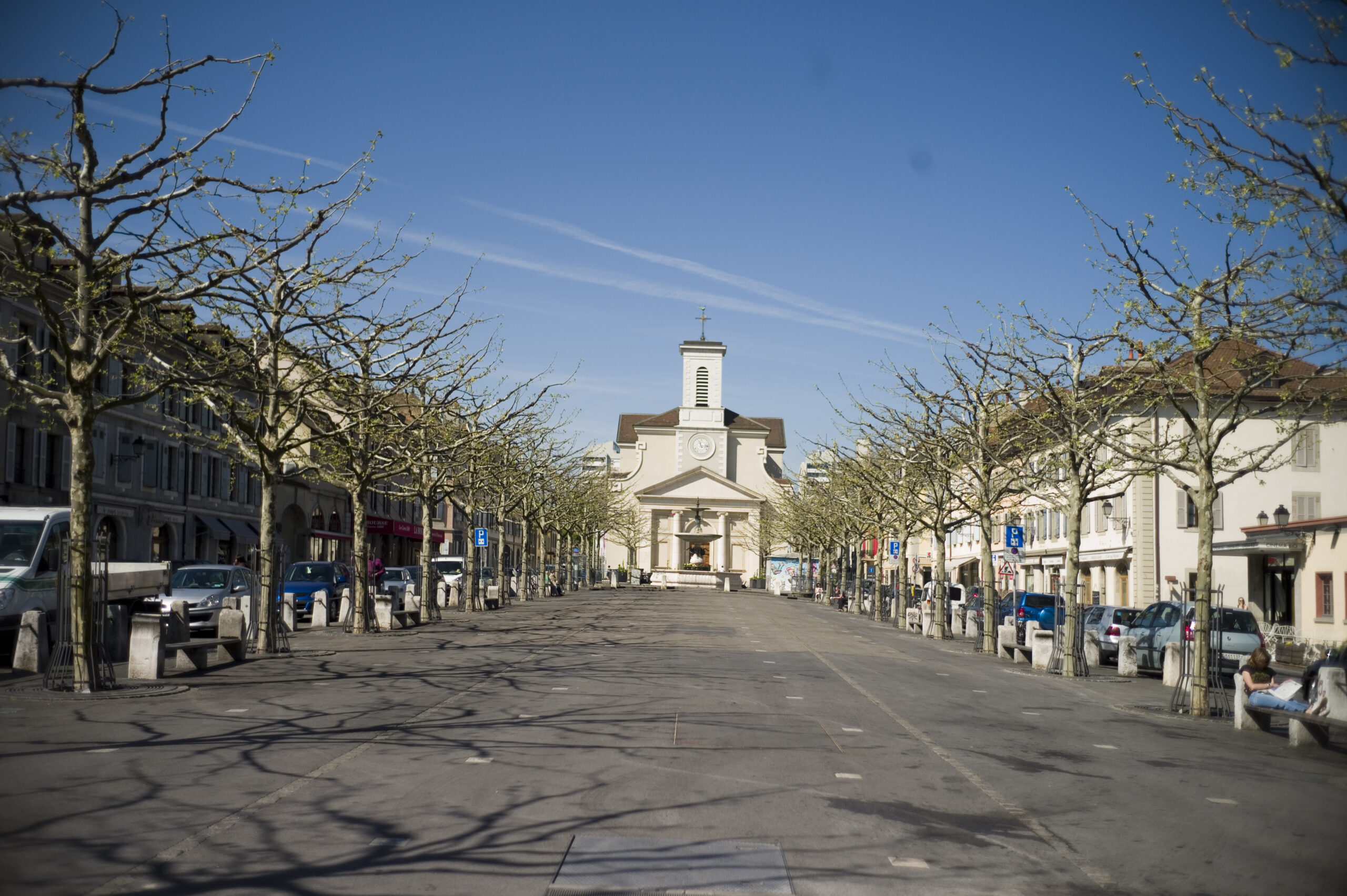 Place du Marché v Carouge s alejí platanů, nízkými domy po stranách a bílým kostelem Sainte-Croix na konci náměstí