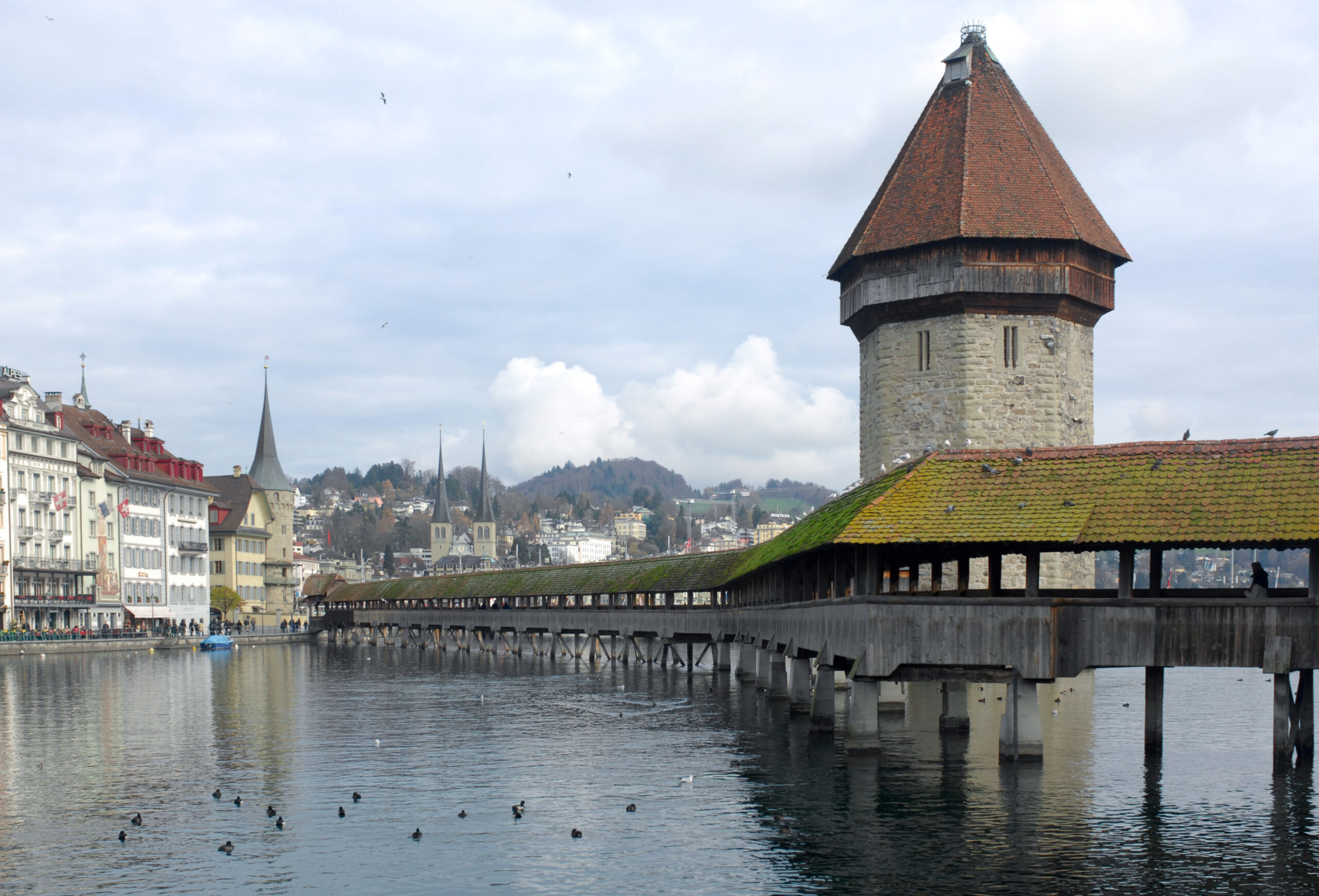 Krytý dřevěný most Kapellbrücke s osmibokou kamennou věží Wasserturm na řece Reuss v Lucernu, vlevo domy na nábřeží a věže kostela Hofkirche
