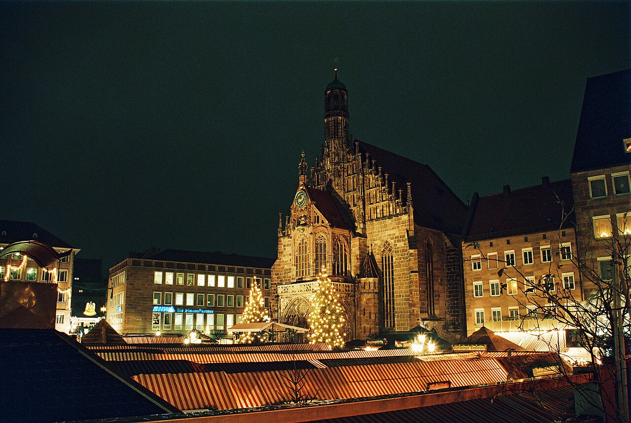 The illuminated Frauenkirche above the stall roofs of the Christkindlesmarkt in Nuremberg at night, with Christmas trees in front of the church