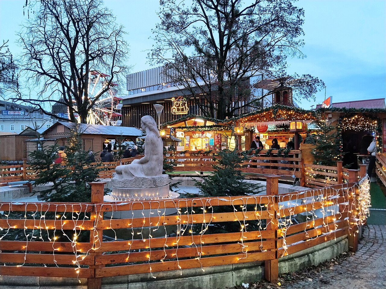 Vánoční trh ve Volksgartenu v Linci: dřevěné stánky se světýlky kolem kašny se sochou, vzadu vyhlídkové kolo a budova Musiktheateru