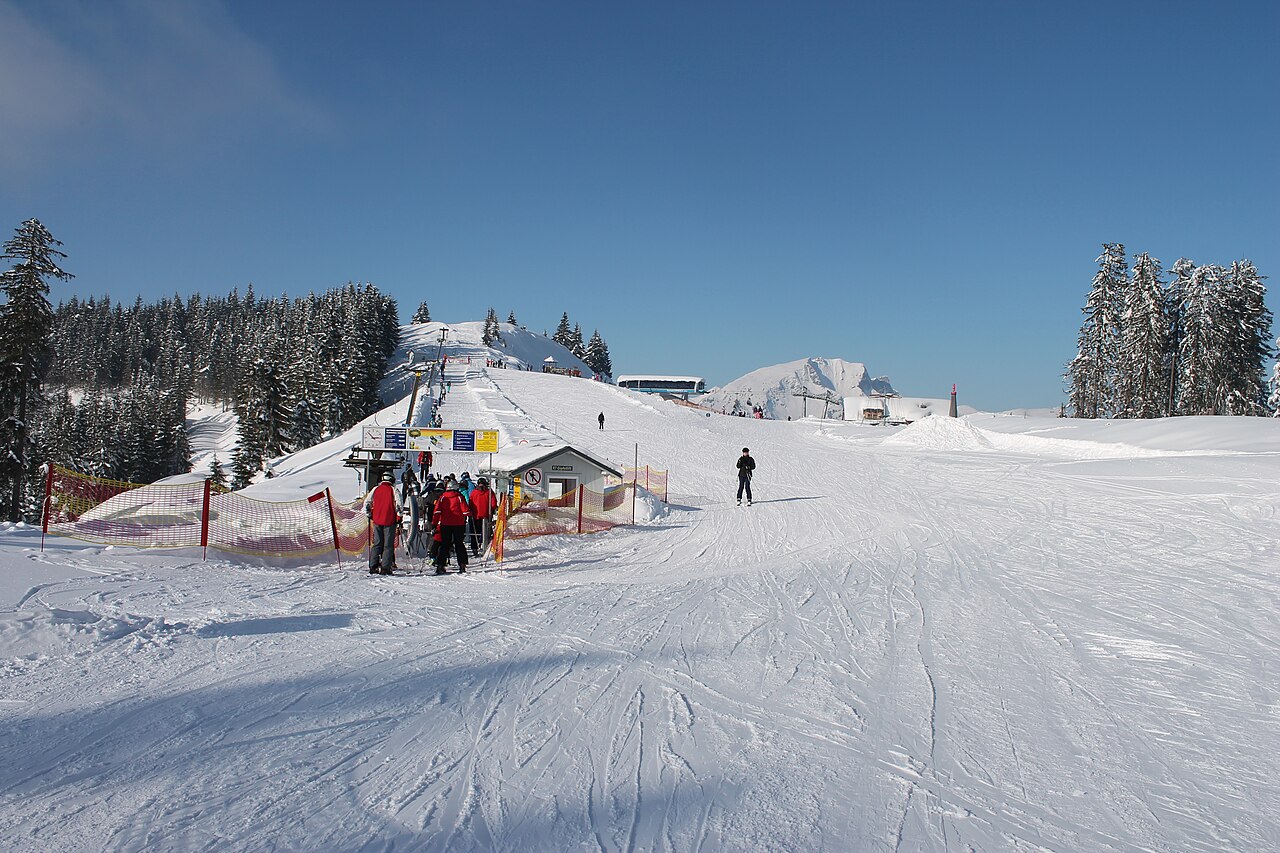 Vlek pod vrcholem Hornspitz v areálu Dachstein West, lyžaři u nástupu a široká upravená sjezdovka pod modrou oblohou