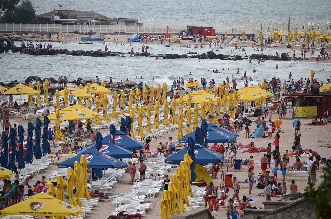 Plná pláž v Eforie Nord se slunečníky, lehátky a kamennými vlnolamy v moři