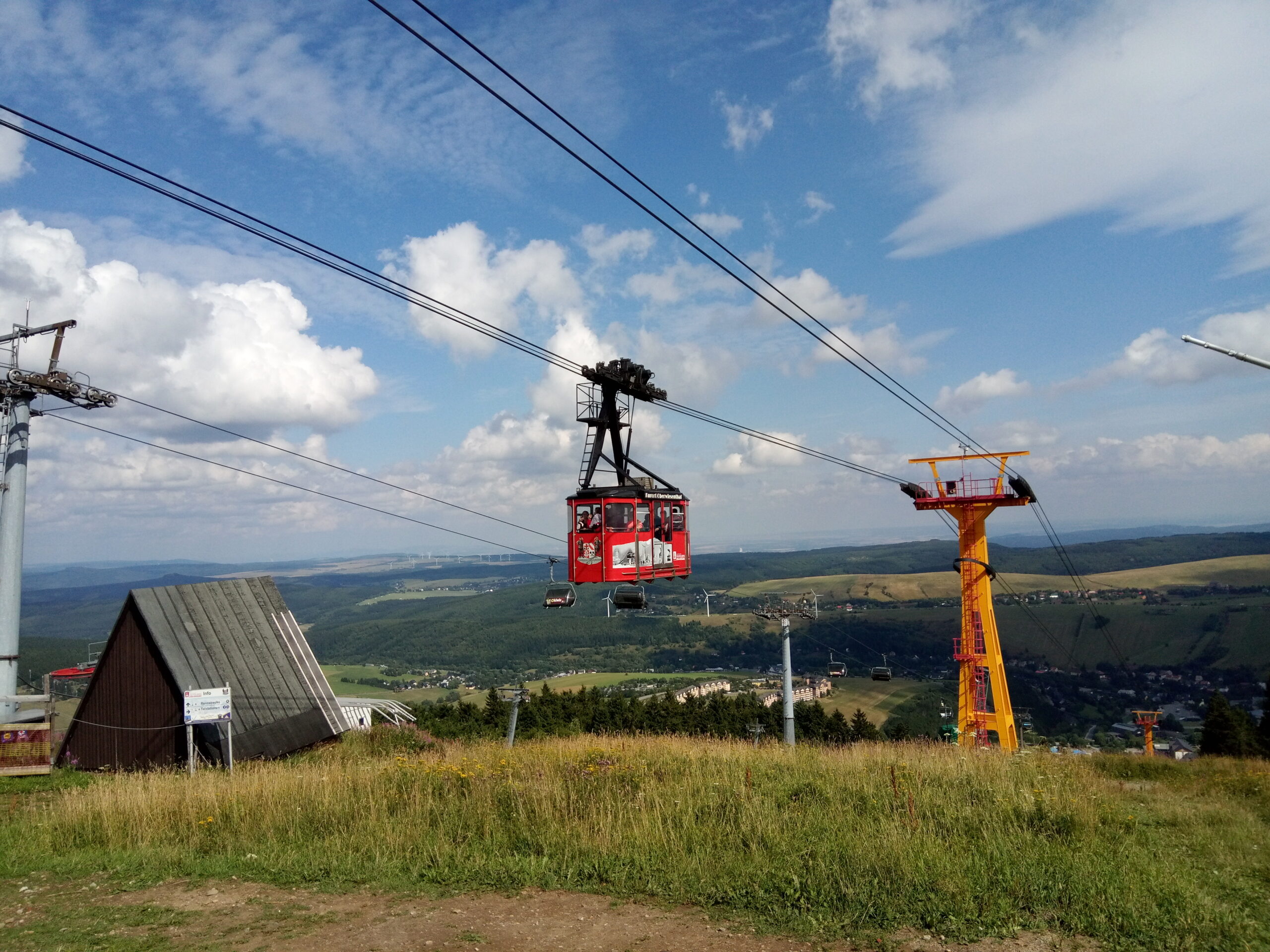 Červená kabina Fichtelberg-Schwebebahn na laně pod vrcholem Fichtelbergu, žlutý stožár a pod ním Oberwiesenthal a zelené kopce