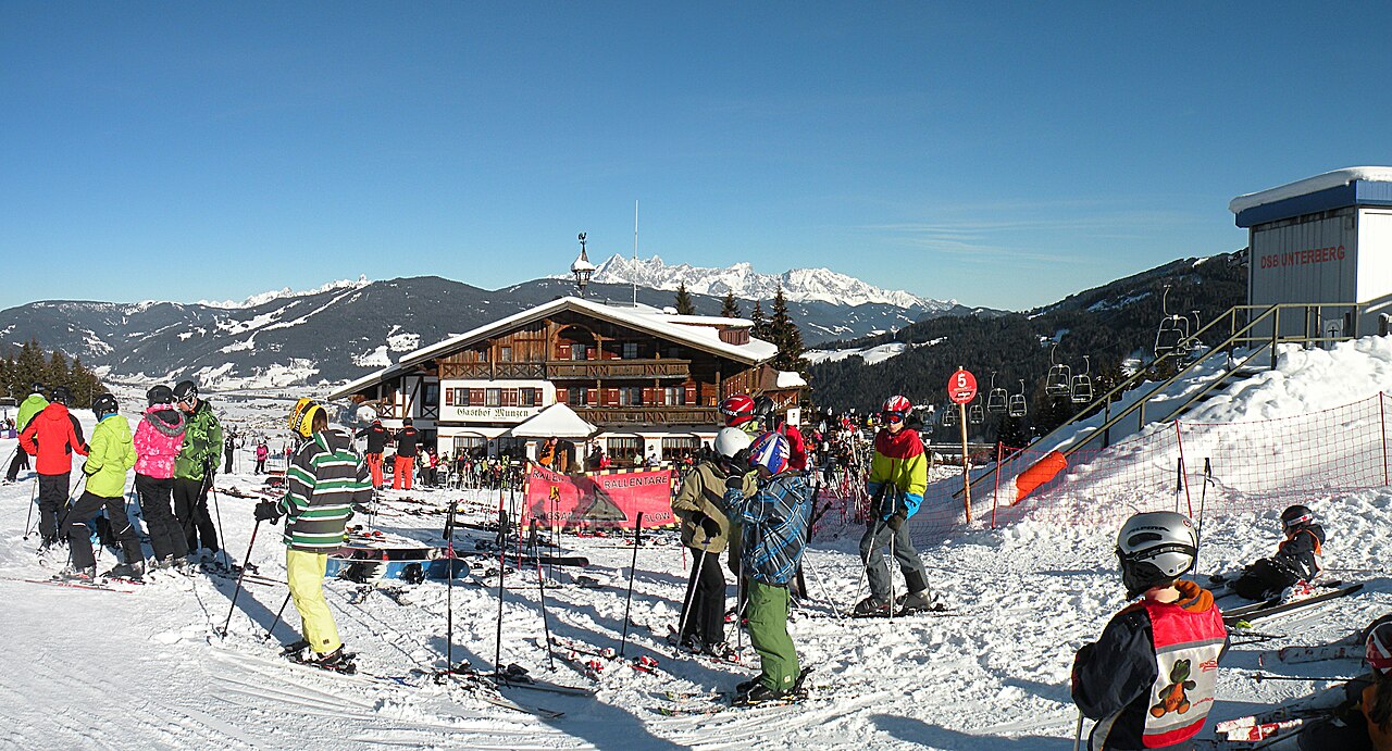 Lyžaři před horskou chatou Gasthof Munzen na sjezdovce ve Flachau, za ní sedačková lanovka a v dálce zasněžený masiv Dachsteinu