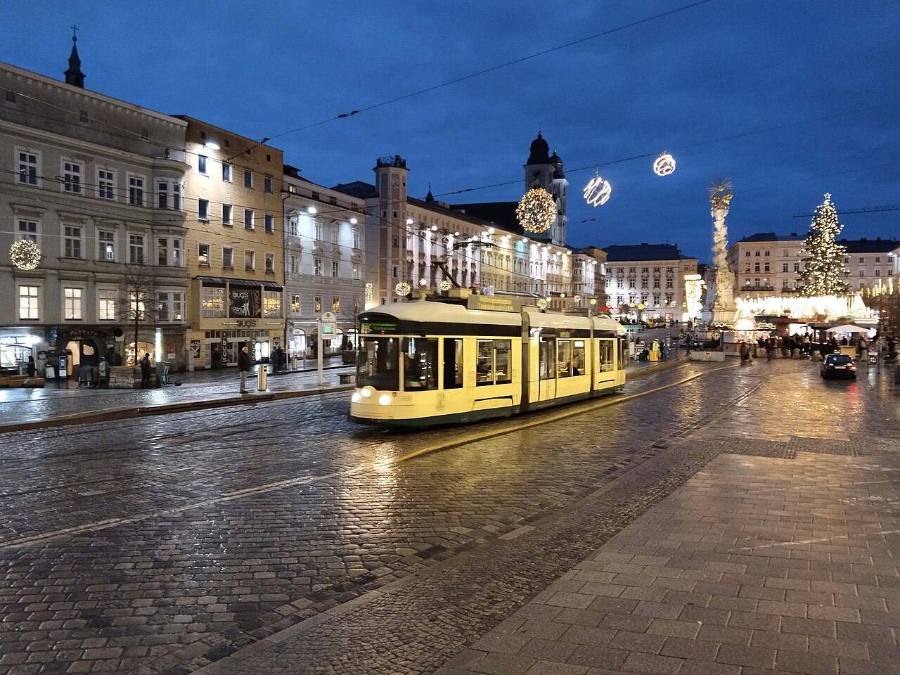 Tramvaj Pöstlingbergbahn na mokré dlažbě Hauptplatzu v Linci v adventu, v pozadí sloup Nejsvětější Trojice a rozsvícený vánoční strom
