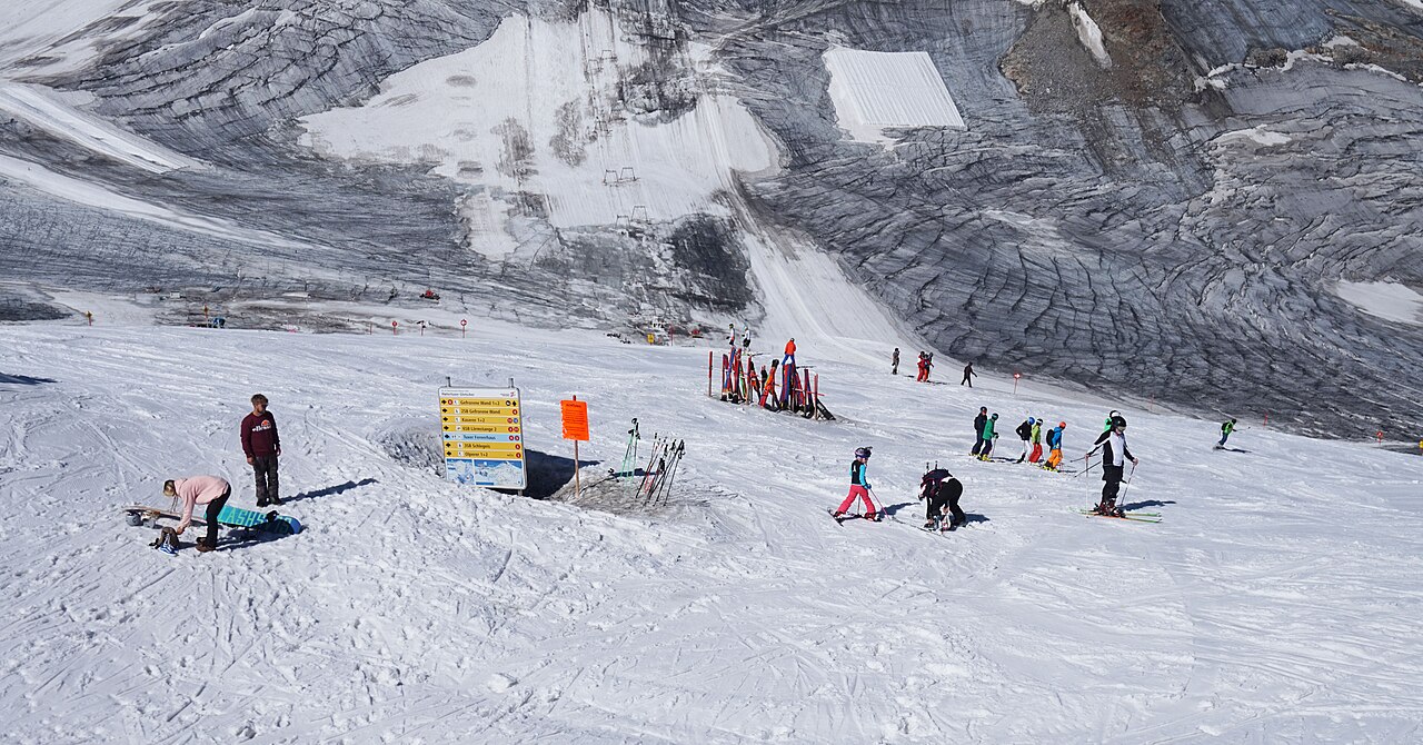 Lyžaři a snowboardisté na upravené sjezdovce ledovce Hintertux, za nimi rozpraskaný led ledovce a ukazatel tratí