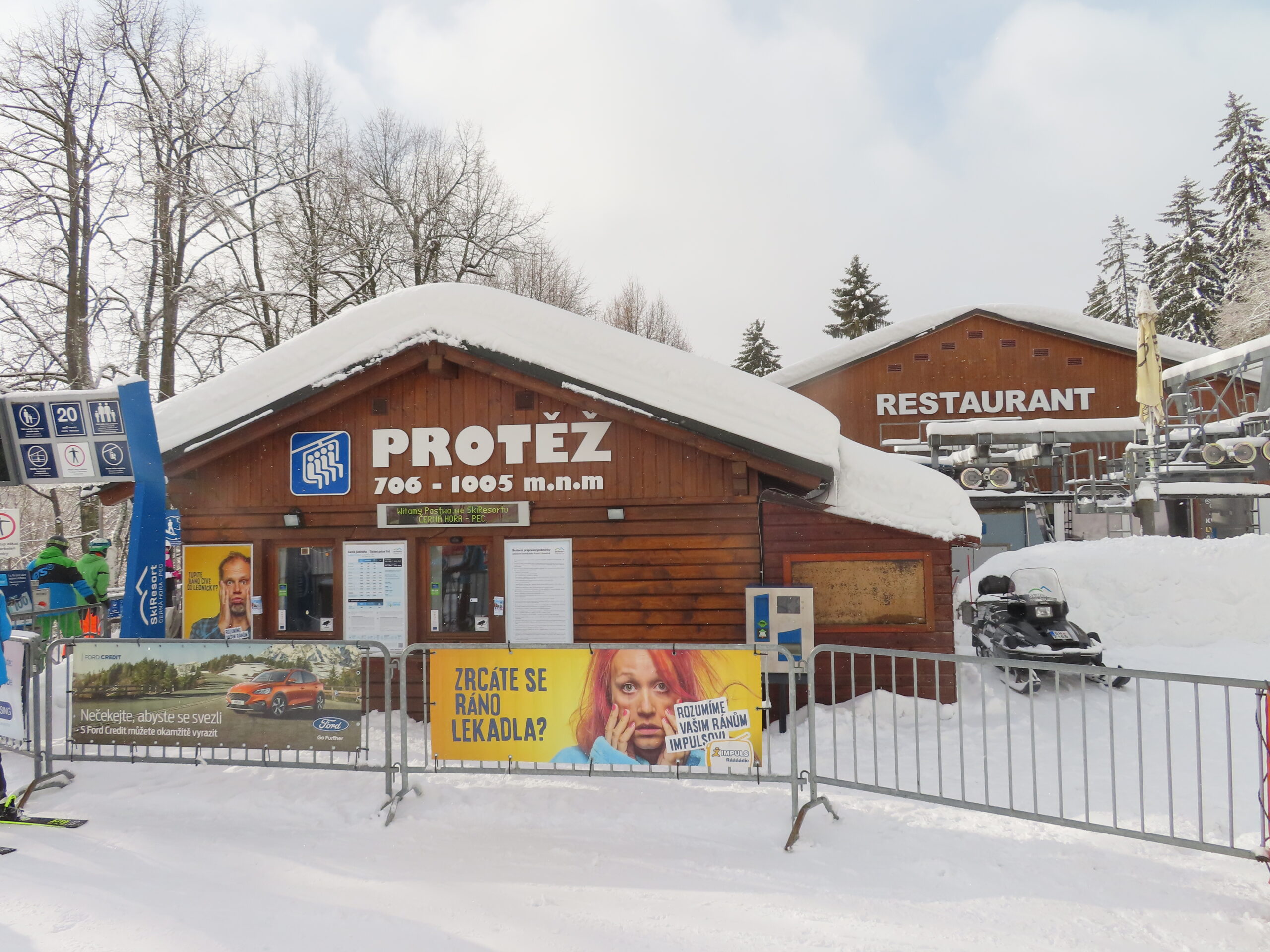 Zasněžená dřevěná budova dolní stanice lanovky Protěž s nápisem 706 až 1005 m n. m., vedle restaurace a nástupiště sedačkové lanovky