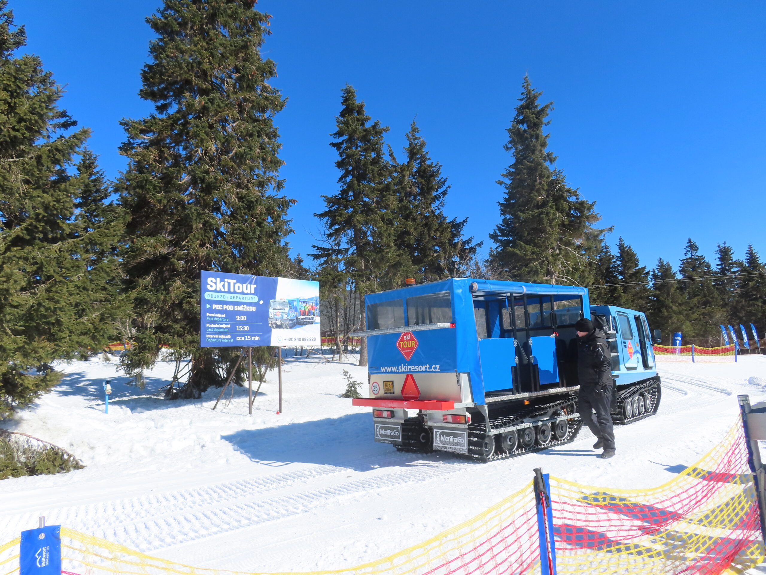 Modrá pásová rolba SkiTour na zasněžené pláni na Černé hoře u cedule s odjezdy do Pece pod Sněžkou, kolem vysoké smrky