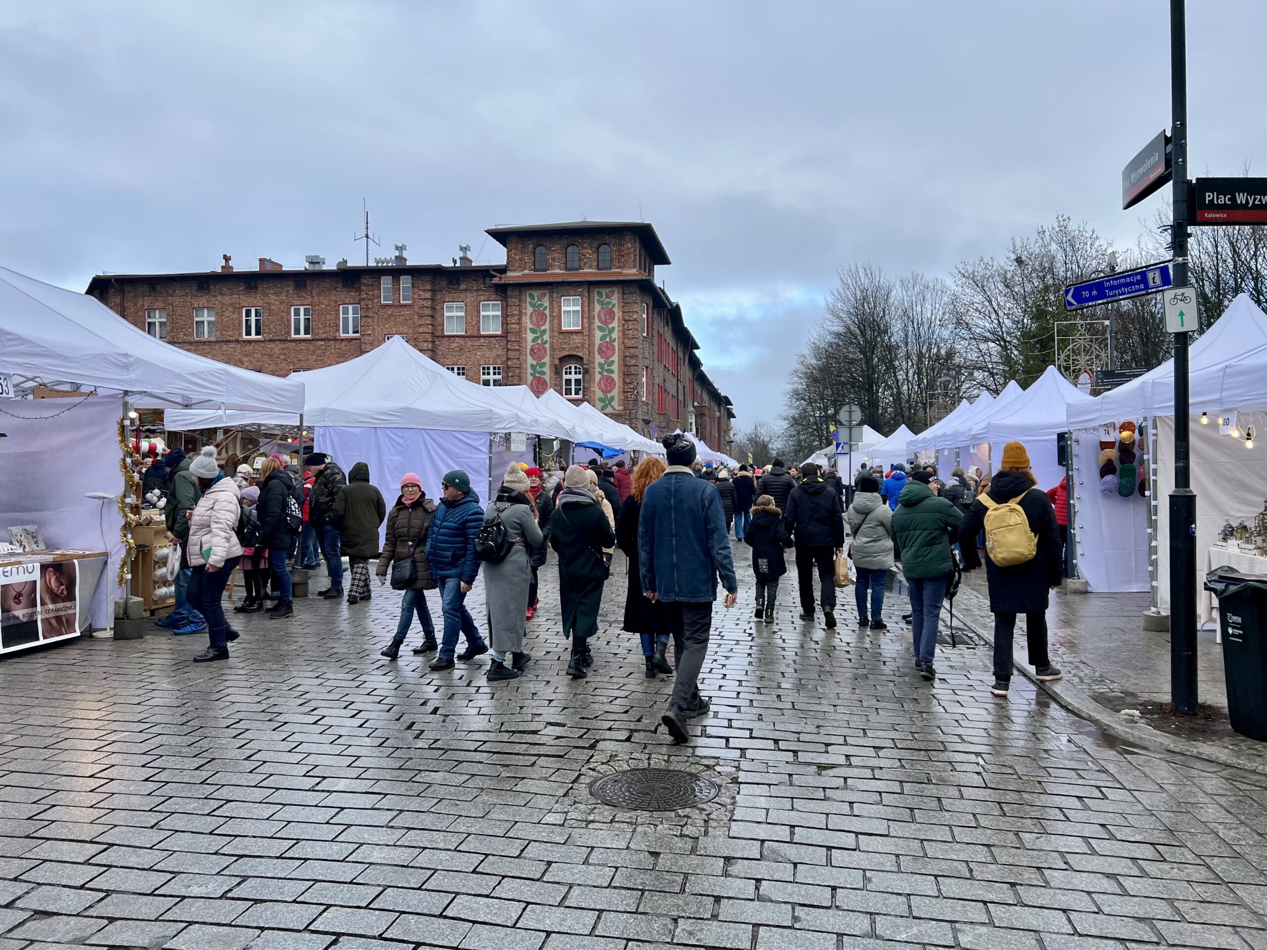 Bílé stany vánočního jarmarku a lidé v zimních bundách na placu Wyzwolenia v Nikiszowci, v pozadí cihlový dům horníků