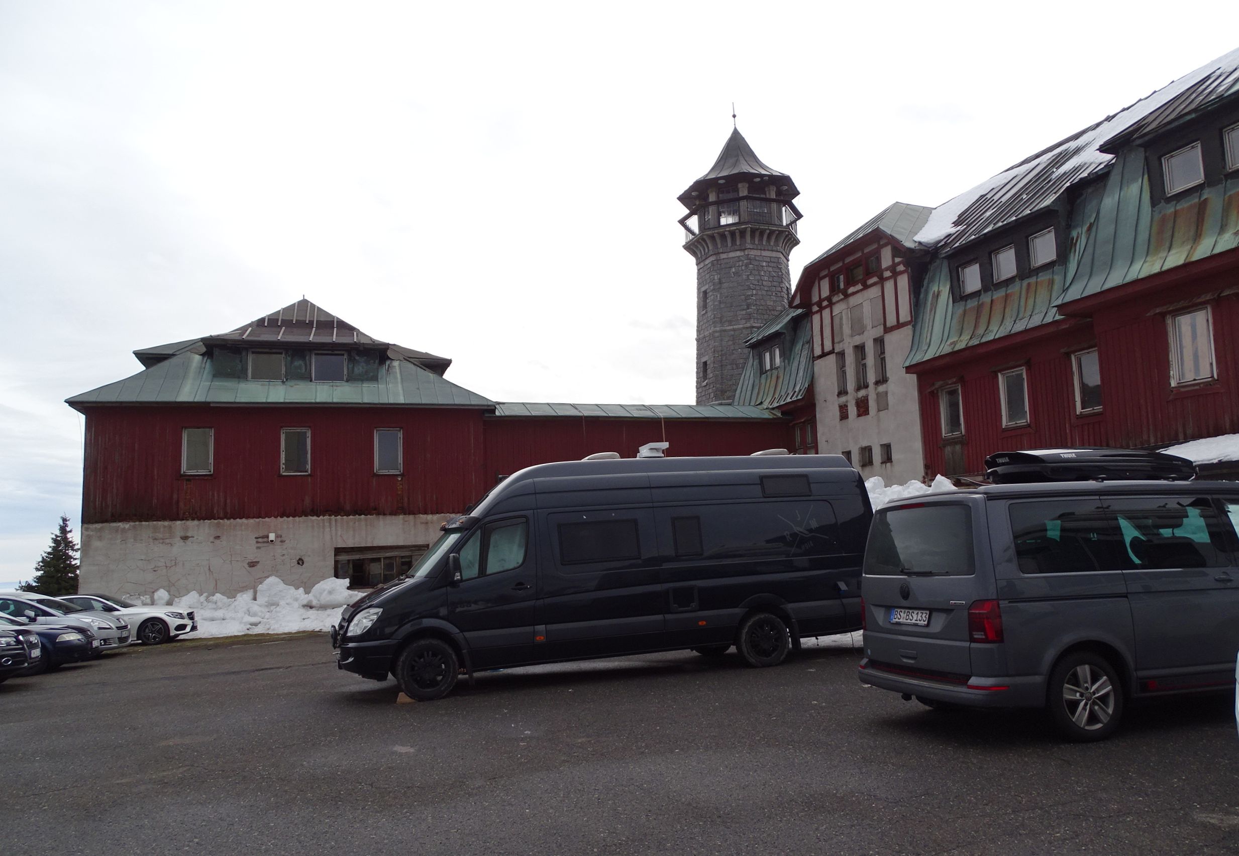 Červeně natřený dřevěný horský hotel na vrcholu Klínovce s kamennou rozhlednou, před ním zaparkovaná auta a zbytky sněhu