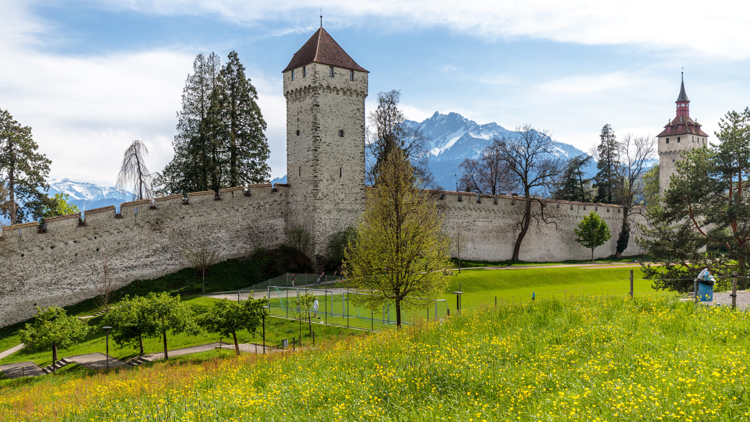 Středověká hradba Museggmauer v Lucernu s kamennými věžemi Wachtturm a Zytturm nad rozkvetlou loukou, v pozadí zasněžené hory