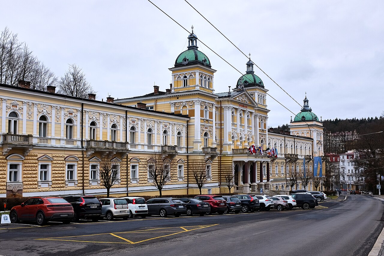 Edificio amarillo neorrenacentista del hotel Nové Lázně en Mariánské Lázně con cúpulas verdes y banderas sobre la entrada