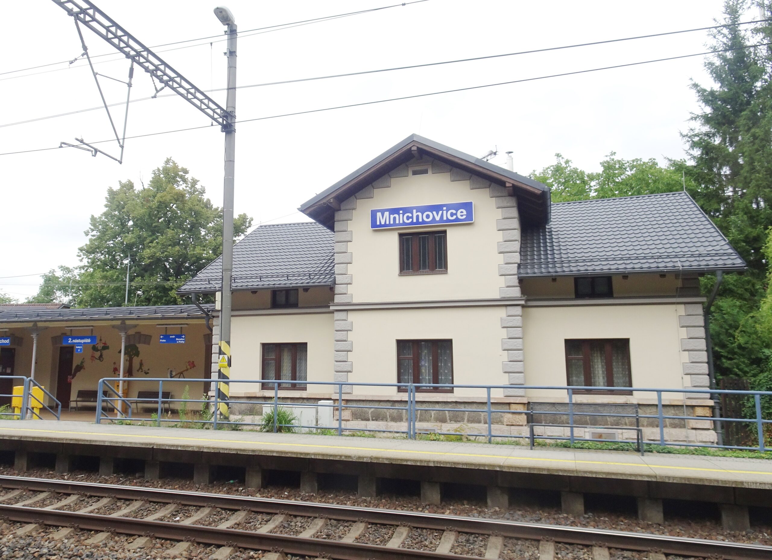 Nádražní budova v Mnichovicích s modrou cedulí s názvem stanice, nástupiště a koleje
