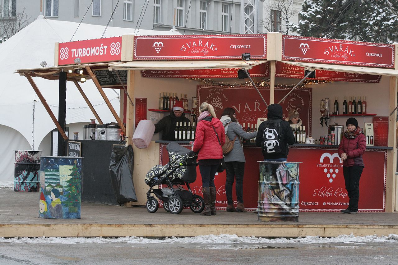 Stragan z napisami Turbomošt i Svařák na jarmarku bożonarodzeniowym na placu Morawskim w Brnie, przed nim ludzie z wózkiem