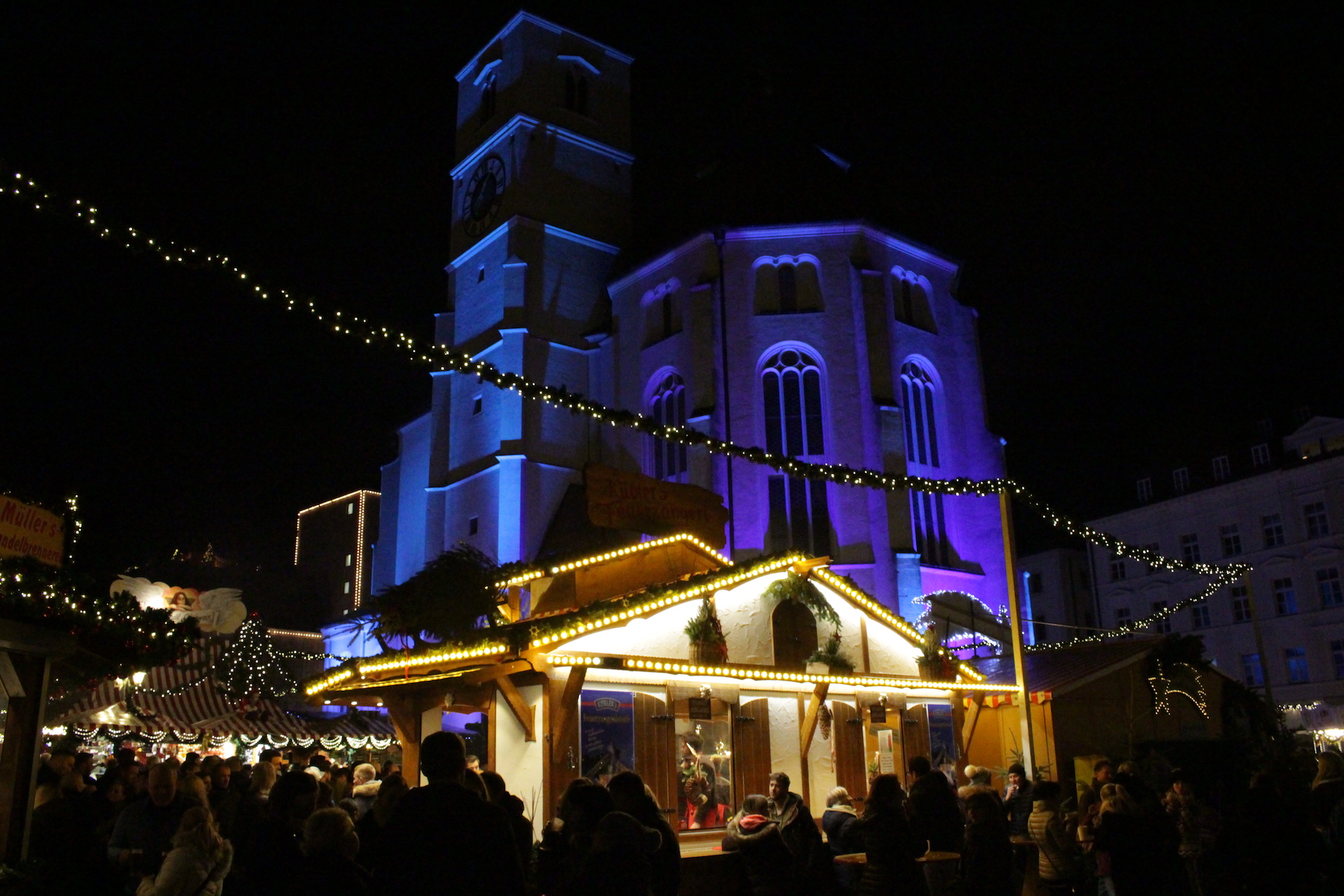 Regensburg vánoční trhy: večerní Christkindlmarkt na Neupfarrplatzu s nasvíceným kostelem Neupfarrkirche a dřevěným stánkem ve světýlkách