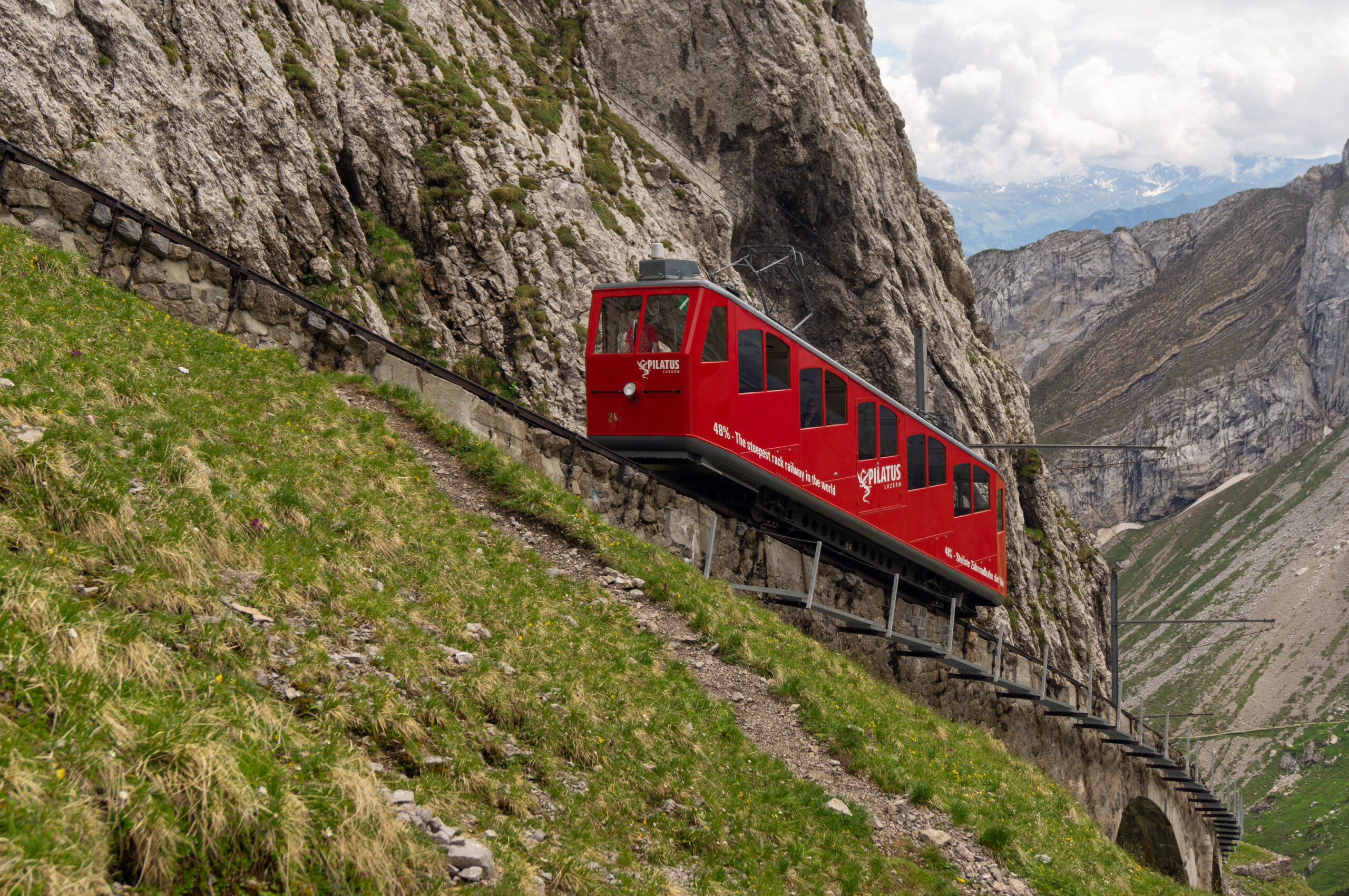 Červený vůz zubačky Pilatusbahn stoupá po strmé trati ve skalní stěně hory Pilatus, v pozadí údolí a vzdálené Alpy