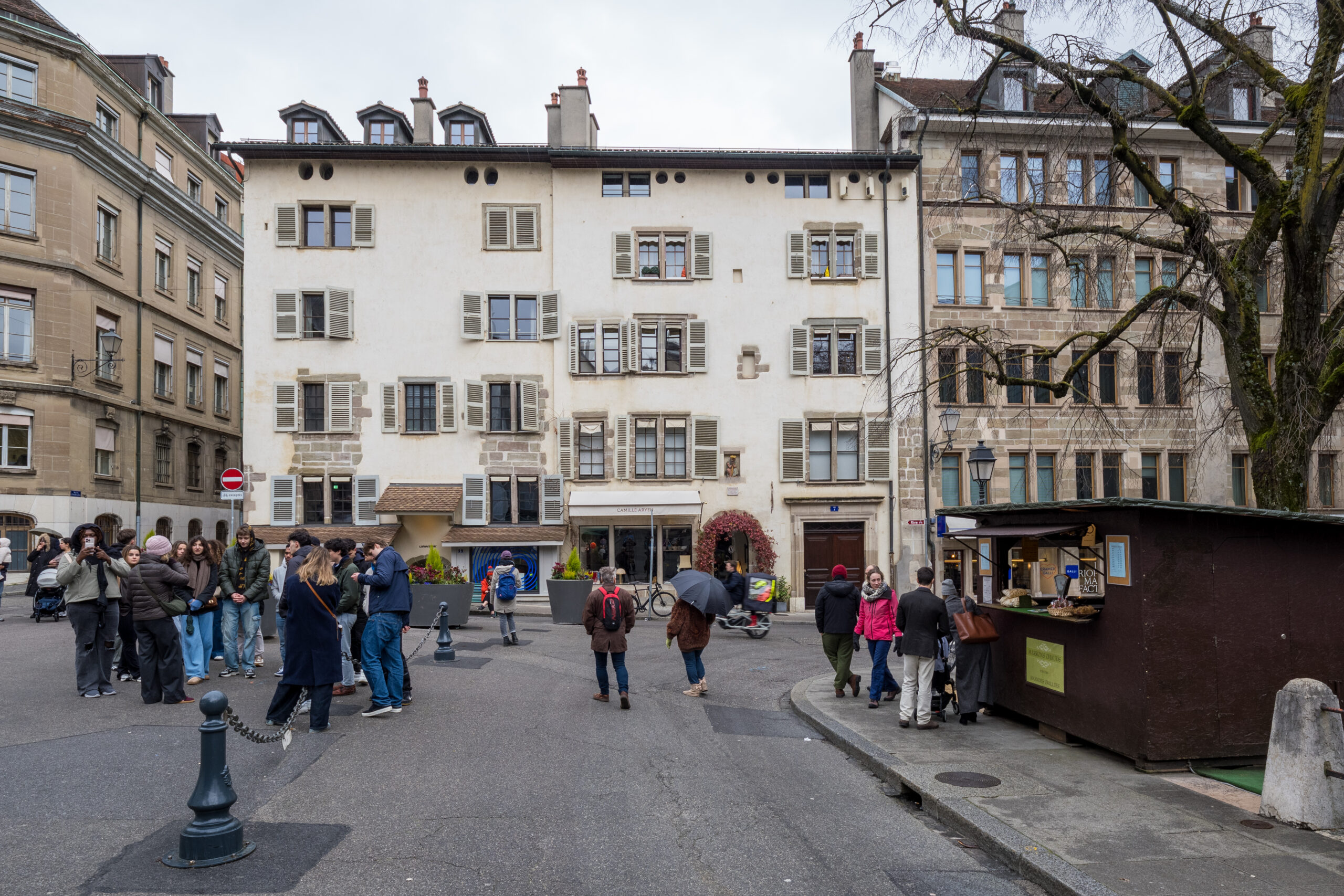 Náměstí Place du Bourg-de-Four ve starém městě Ženevy, světlé měšťanské domy s okenicemi, chodci a stánek