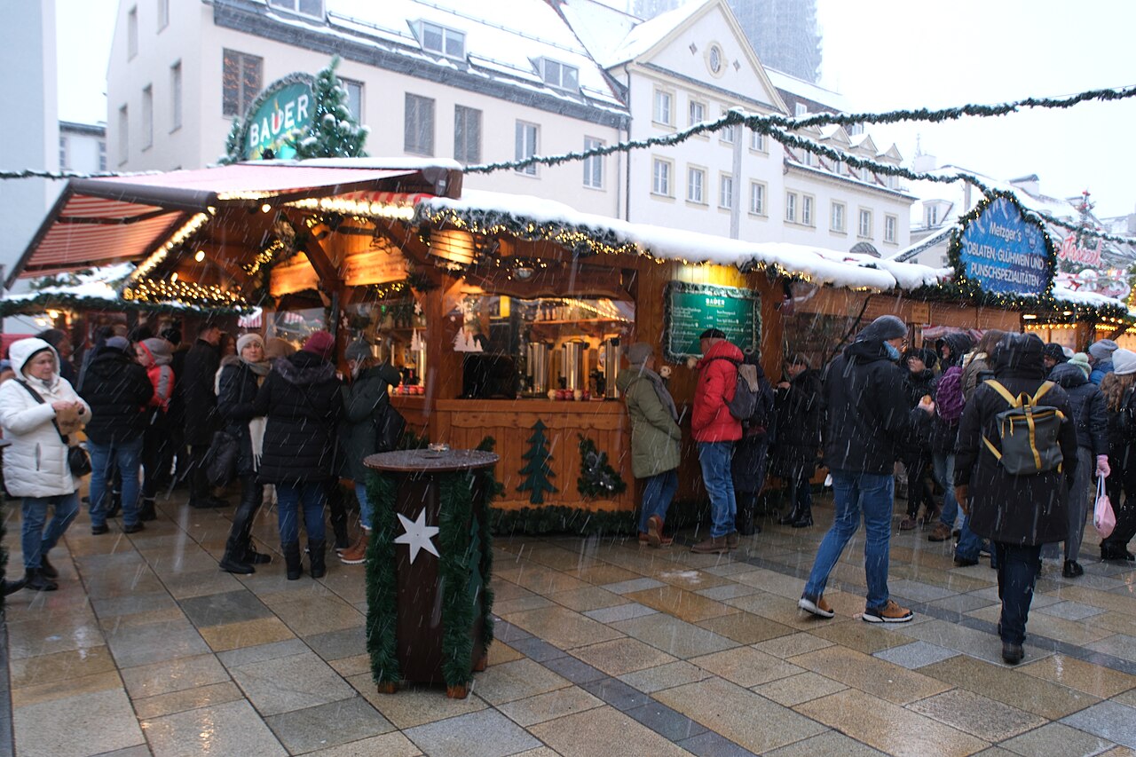 Zasněžené stánky se svařákem a punčem na Christkindlmarktu v Řezně, lidé v zimních bundách při sněžení