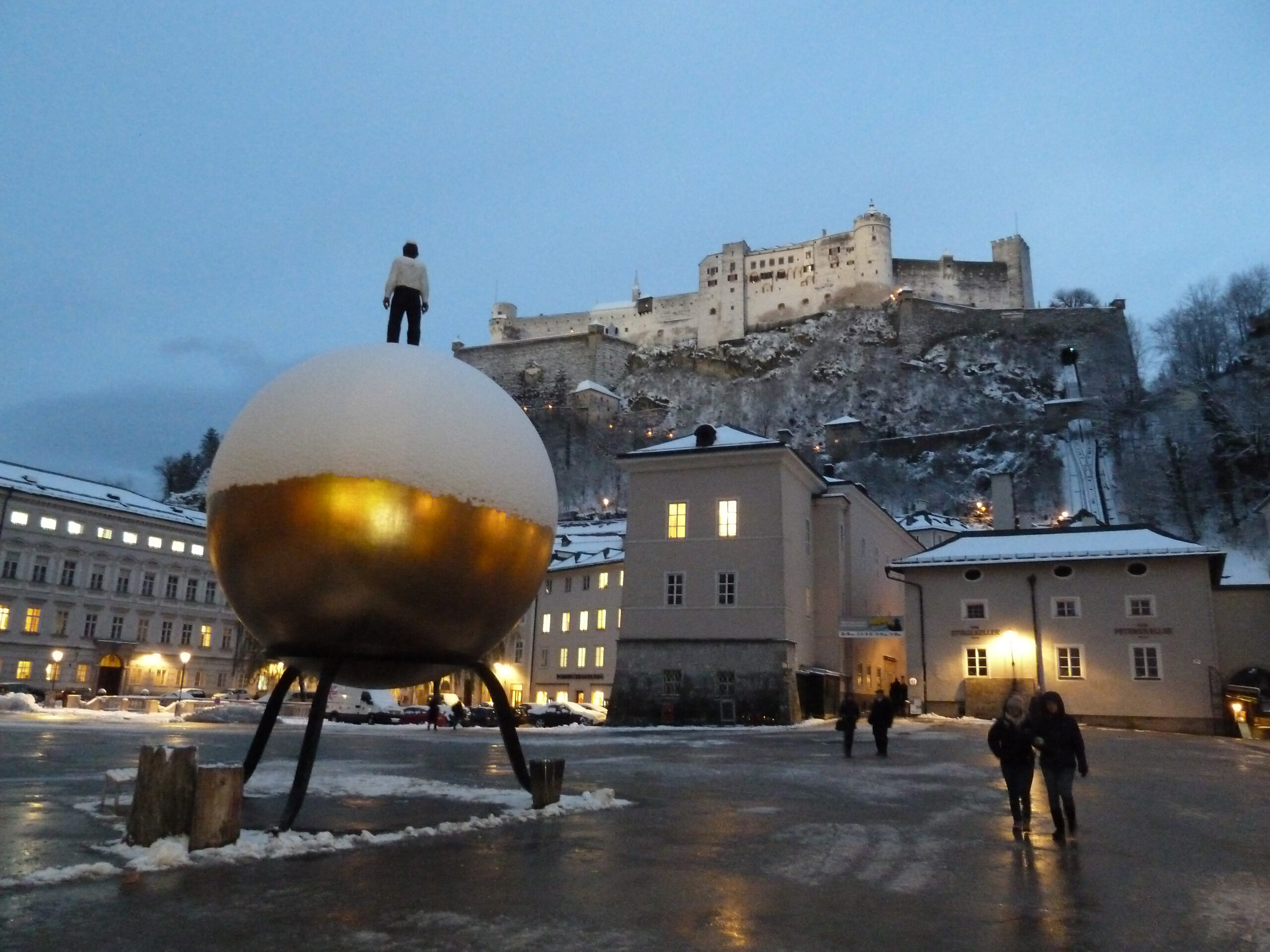 Zasněžený Kapitelplatz v Salcburku za soumraku se zlatou koulí se sochou a pevností Hohensalzburg nad městem