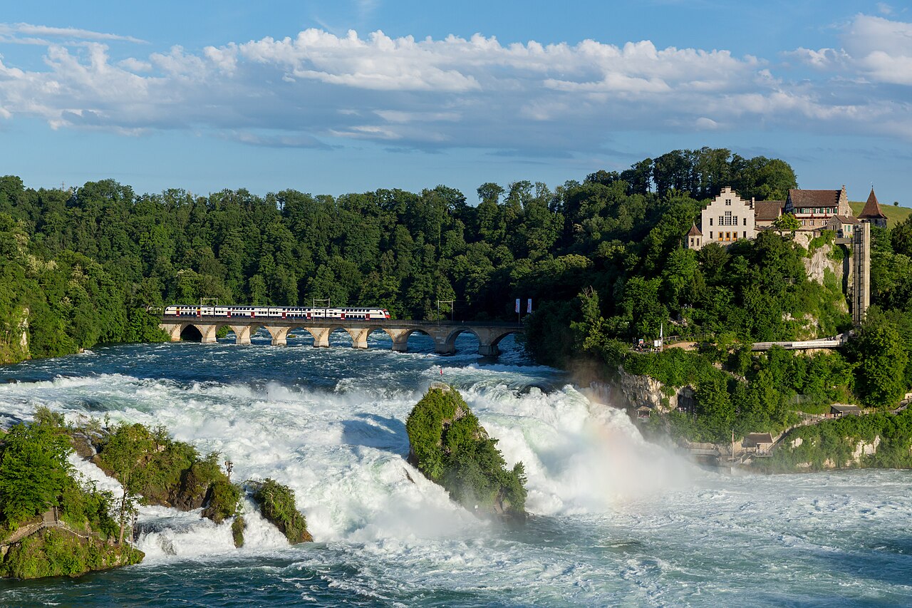 Rýnský vodopád Rheinfall se skálou uprostřed, nad ním železniční most s vlakem S-Bahn a vpravo na útesu zámek Laufen
