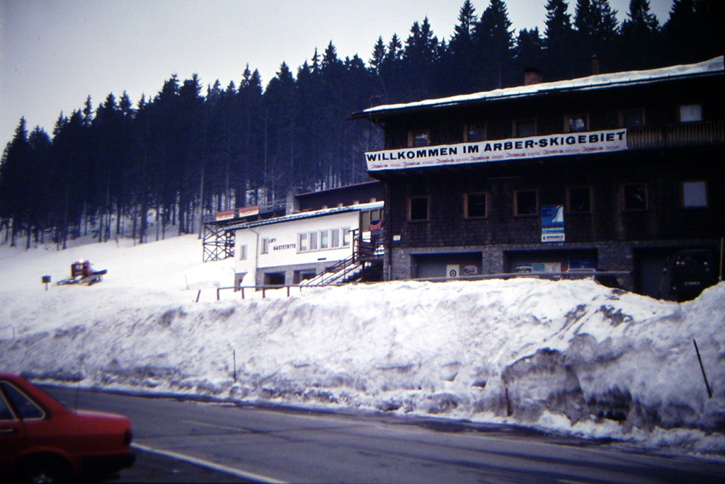 Dřevěná budova s transparentem Willkommen im Arber-Skigebiet a vysoké mantinely sněhu podél silnice v roce 1984