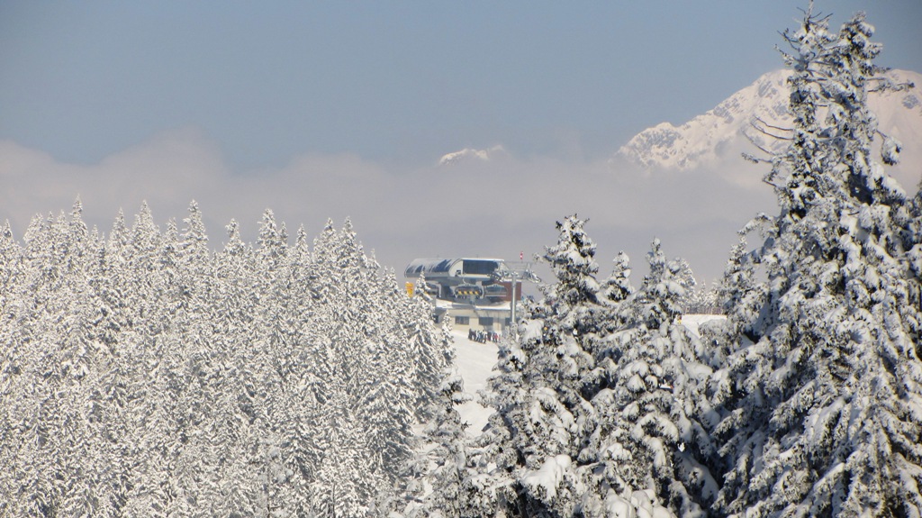 Zasněžené smrky a horní stanice lanovky v oblasti SkiWelt Wilder Kaiser-Brixental, nad mraky vrcholy hor