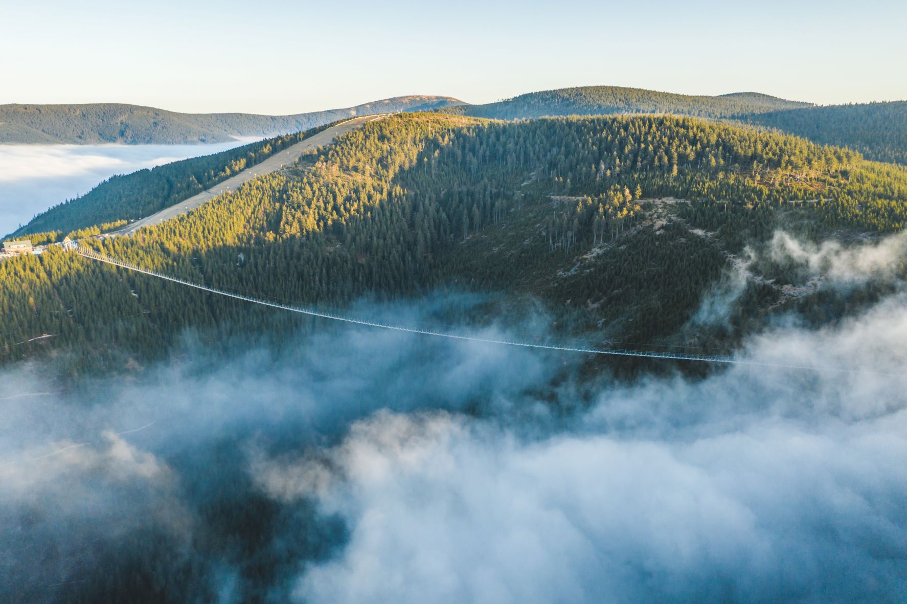 Visutý most Sky Bridge 721 nad zalesněným údolím u Dolní Moravy, údolí zaplněné mlhou, v pozadí hřeben Králického Sněžníku