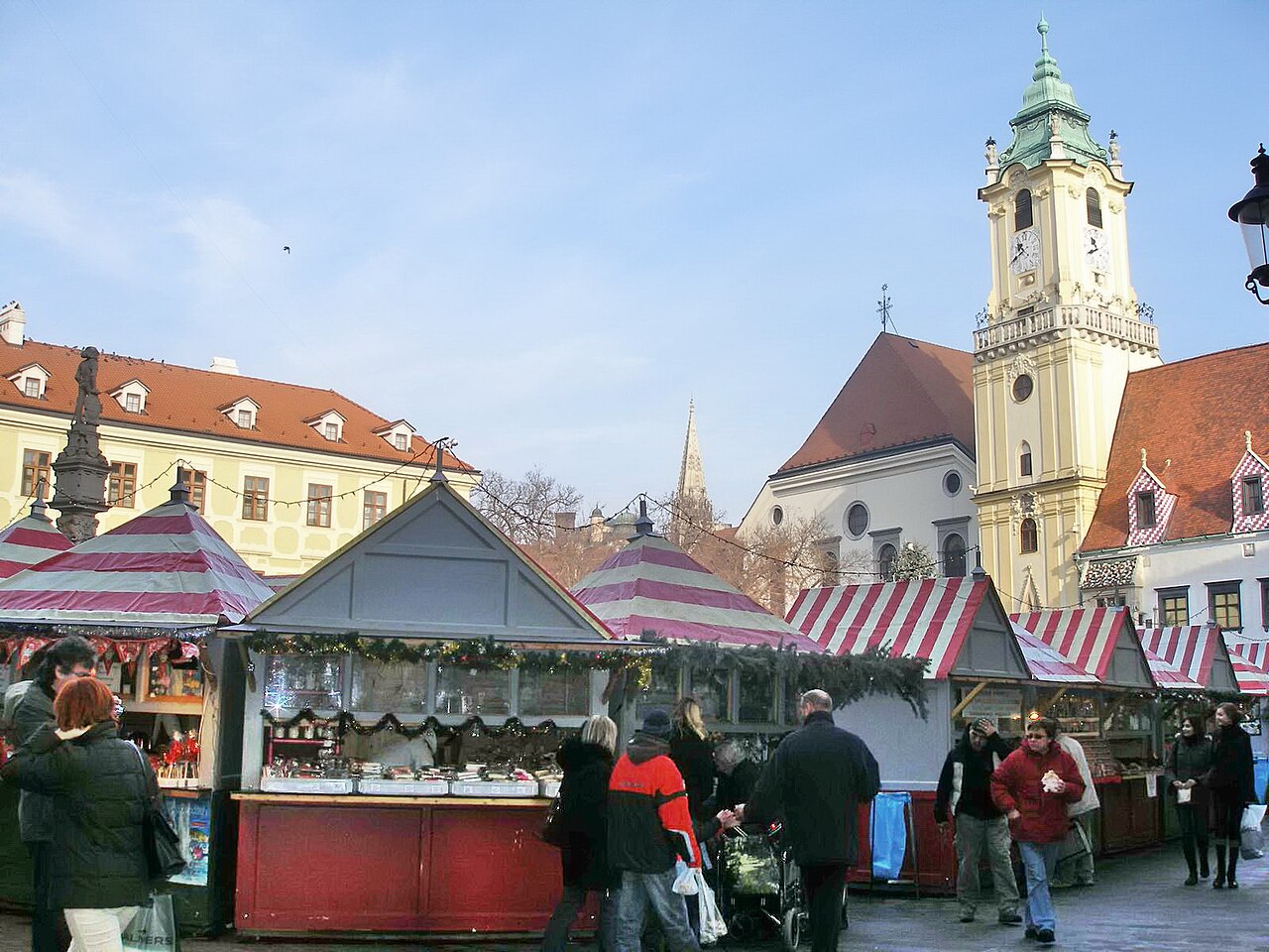 Stánky vánočního trhu s červenobílými střechami na Hlavnom námestí v Bratislavě, vpravo věž Staré radnice