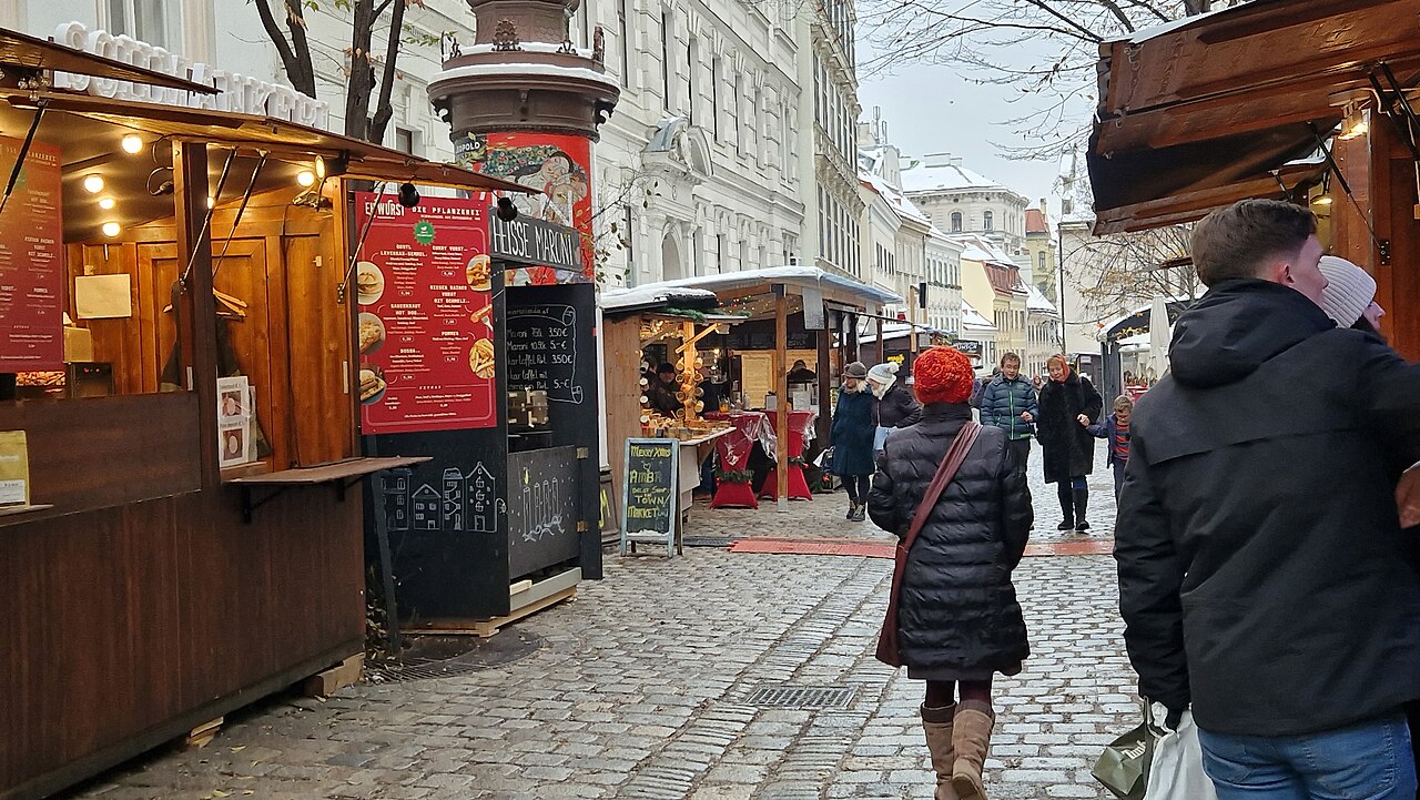 Vianočný trh na Spittelbergu vo Viedni: drevené stánky s jedlom v úzkej dláždenej uličke medzi starými domami, ľudia v zimných bundách