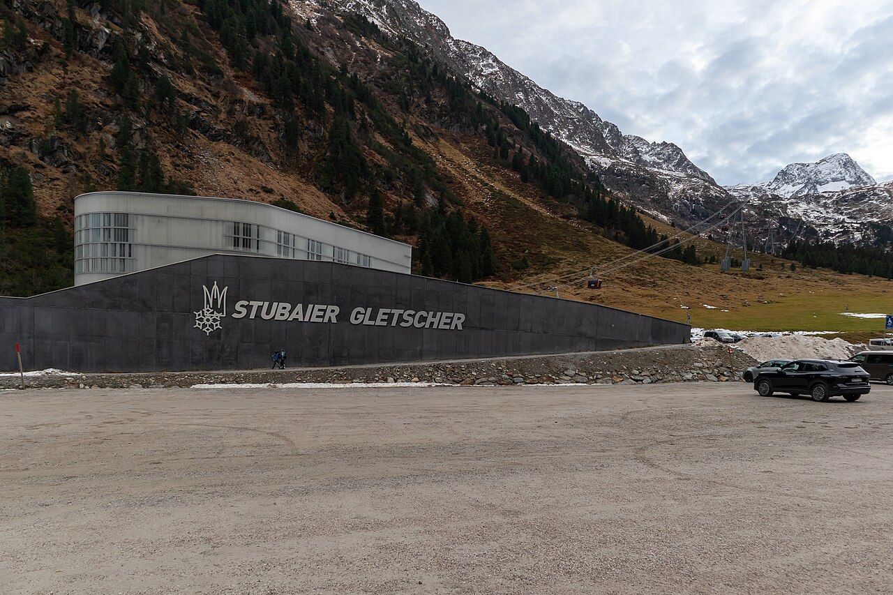 Údolní stanice lanovky Eisgratbahn s nápisem Stubaier Gletscher, parkoviště a kabinky lanovky stoupající do zasněžených hor