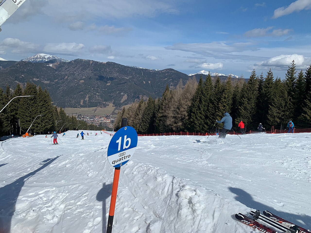 Široká modrá sjezdovka 1b na Stuhlecku s lyžaři, v údolí obec Spital am Semmering a za ní zalesněné kopce se zasněženými vrcholy
