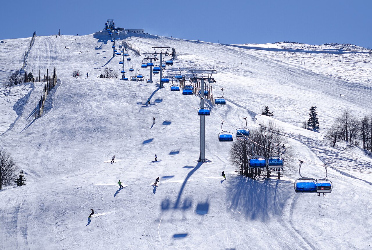 Sedačková lanovka s modrými sedačkami nad širokou sjezdovkou v Uludağu, lyžaři a snowboardisti za slunečného dne