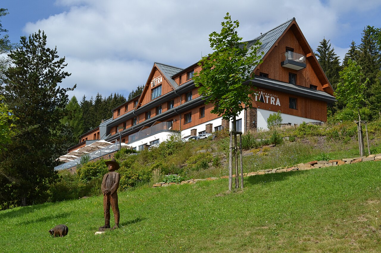 Edificio revestido de madera del Grandhotel Tatra en Velké Karlovice sobre una ladera verde, en primer plano una escultura de madera de un pastor