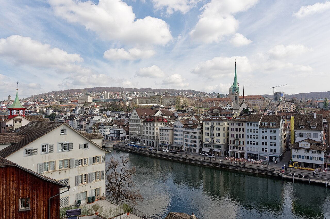 Výhled z Lindenhofu v Curychu na řeku Limmat, nábřeží Limmatquai s měšťanskými domy a zelenou věž kostela Predigerkirche, v pozadí kopec Zürichberg