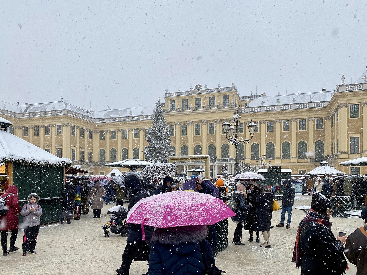 Vianočný trh pred zámkom Schönbrunn počas sneženia, ľudia s dáždnikmi medzi stánkami, vzadu zasnežený vianočný strom a žltá fasáda zámku